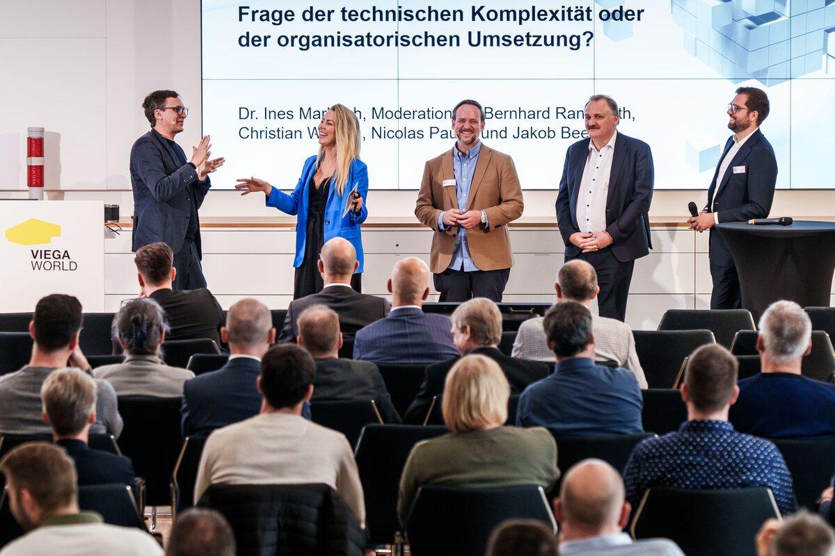 Dr. Ines Marbach im blauen Anzug moderiert eine lebendige Gesprächsrunde vor Publikum