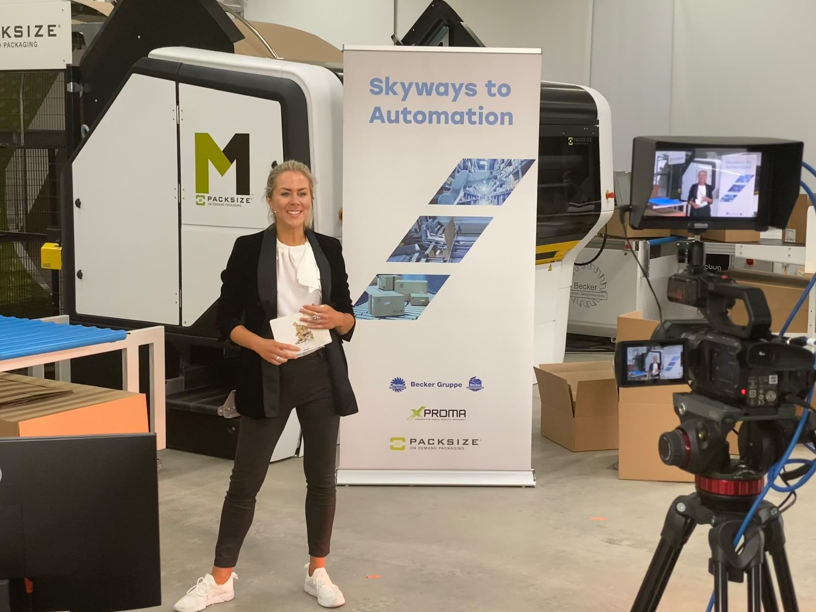 Dr. Ines Marbach bei einer Videoproduktion vor einer Packsize-Verpackungsmaschine, mit einer Kamera im Vordergrund.